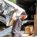 A CORT Furniture Rental delivery professional carefully carrying a large, wrapped furniture piece on his shoulder, with boxes and a CORT truck in the background, showcasing the hands-nature of CORT White Glove service.