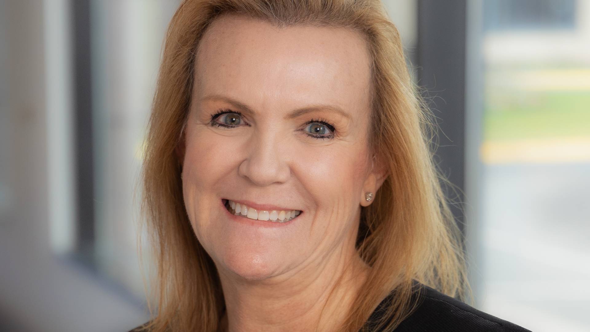 Portrait of Paula Newell, a professional woman with blonde hair and a warm smile, wearing a dark suit, standing in a well-lit indoor setting.