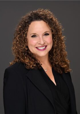 Professional headshot of Angi Pusateri-Downey, smiling warmly, with curly brown hair, wearing a black blazer and a simple necklace against a neutral gray background.