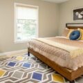 A sunny dorm room with a bed, dresser, desk, and rug.