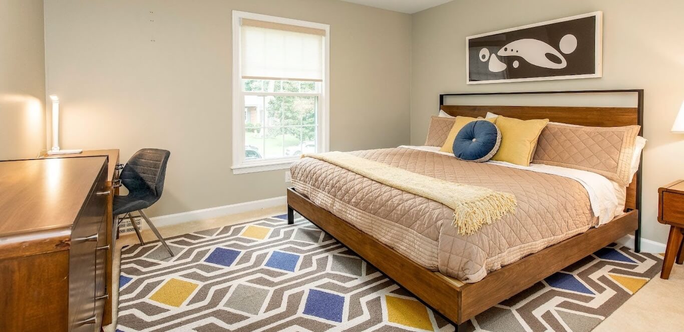 A sunny dorm room with a bed, dresser, desk, and rug.