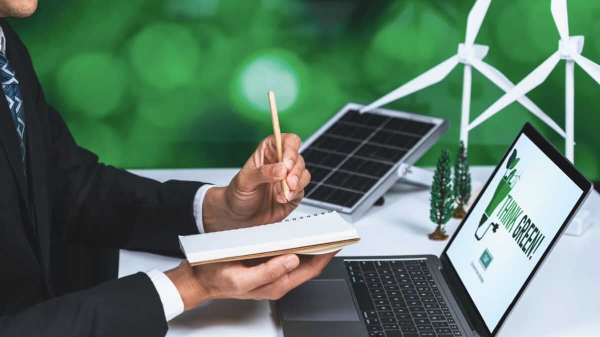 A professional in a suit holding a notebook and pencil, working at a desk with a laptop displaying a "Think Green" message, a miniature wind turbine, solar panel, and small trees in the background, symbolizing renewable energy and sustainability.