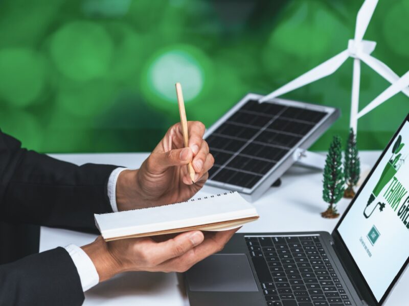 A professional in a suit holding a notebook and pencil, working at a desk with a laptop displaying a "Think Green" message, a miniature wind turbine, solar panel, and small trees in the background, symbolizing renewable energy and sustainability.