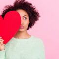 A woman playfully holds a red heart-shaped cutout in front of her face against a pink background.