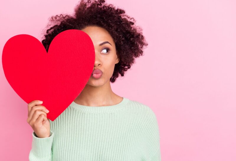 A woman playfully holds a red heart-shaped cutout in front of her face against a pink background.