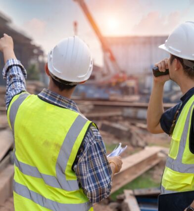 two construction project managers in safety gear discussing site progress at a busy construction site.