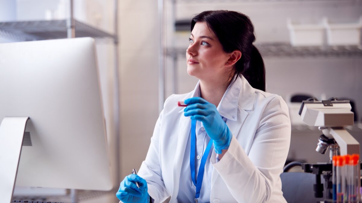 Traveling phlebotomist after learning how to be a travel phlebotomist sitting in a lab with gloves on holding a vial in front of a computer looking off into the distance wistfully.