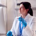 Traveling phlebotomist after learning how to be a travel phlebotomist sitting in a lab with gloves on holding a vial in front of a computer looking off into the distance wistfully.