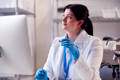 Traveling phlebotomist after learning how to be a travel phlebotomist sitting in a lab with gloves on holding a vial in front of a computer looking off into the distance wistfully.