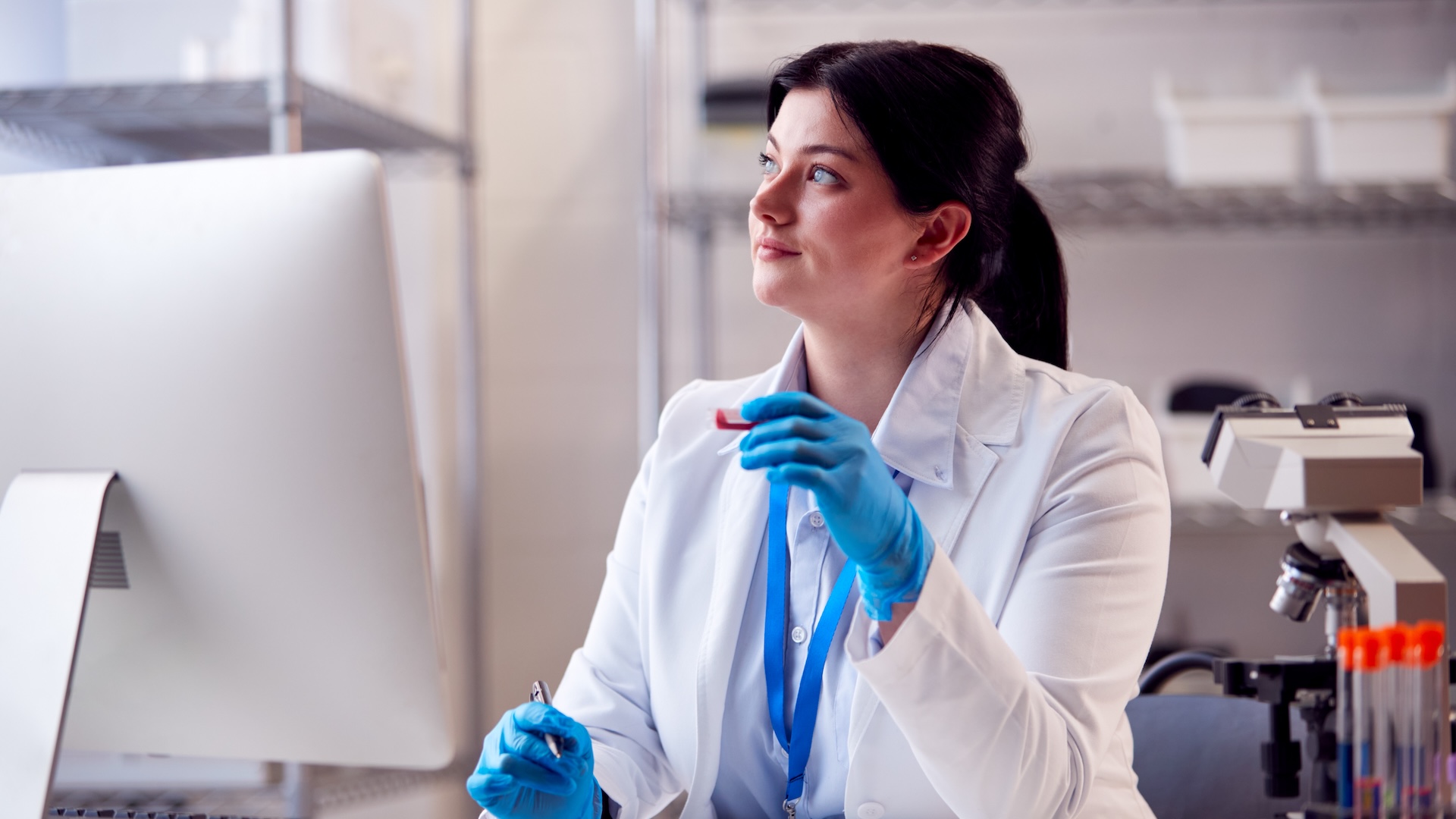 Traveling phlebotomist after learning how to be a travel phlebotomist sitting in a lab with gloves on holding a vial in front of a computer looking off into the distance wistfully.