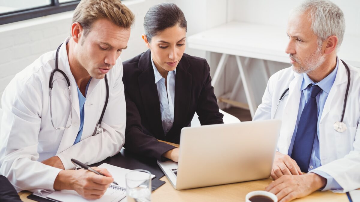 Three healthcare professionals, including two doctors and administrator, collaborate on workspace planning using a laptop in a clinical office seating.