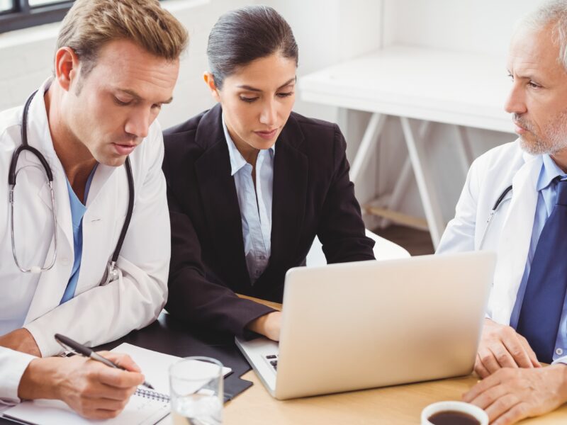 Three healthcare professionals, including two doctors and administrator, collaborate on workspace planning using a laptop in a clinical office seating.